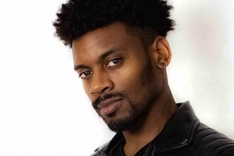 A man with tightly coiled black hair and a goatee looks at the camera, his head angled slightly down. He is wearing a black t shirt, black leather jacket, and a small black earring stud.