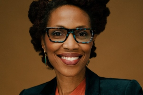A woman with blue and black glasses smiles at the camera. She is wearing teal pendant earrings, red lipstick, a dark green suit jacket, and an orange shirt. The background behind her is a flat brown color.