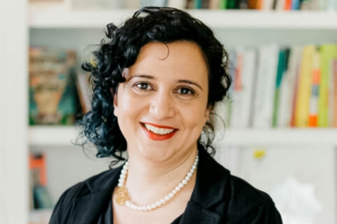 A woman with short curly black hair smiles at the camera. She is wearing a pearl necklace, a black suit jacket and black shirt, and red lipstick. The background behind her are of white bookshelves with different books of varying bright colors.