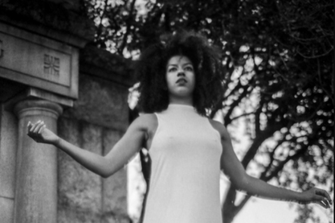 A black and white image of a woman with an afro hairstyle and a white dress, with her arms out to her sides as she balances on ballerina toes. She appears to be in a garden, where behind her is a large stone building, and a large clay pot. She is surrounded by plants and there is a large tree behind the stone building.