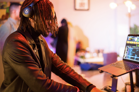 A Dj with short dreads wears Beats Headphones and a black leather jacket as he adjusts a DJ table set, his laptop computer propped up on a stand in front of him, displaying different sound samples. Behind it is a cactus plant. The blurred background behind him is a room with two other people whose faces cannot be seen. In the room is a table with items on top, and a couple of lamps, a picture frame.