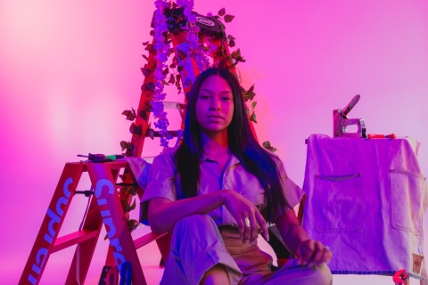 A long haired woman dressed in a white shirt tucked into brown pants sits cross legged in front of two step ladders labelled "Supreme". To her left are wood working tools like a stapler and a hot glue gun, littered on top of a small work table covered with an apron, and on the floor around her legs. The entire image is colored in purple and pink hues.