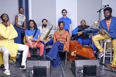 Mwenso and The Shakes, comprised of nine members, including Michael Mwenso, in the yellow on the far left. The band sit in a soundproof room with all of their instruments and speakers.