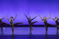 Against a vibrant purple background, five femail dancers balance on the floor on one knee their other leg extened to the side and their arms reaching upwards