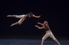 two male dancer wearing black tops and white pants. One man seems to fly through the air to the arms of the other.