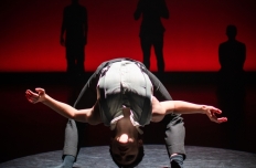 Against a blood red background there are shadowy figures. In the foreground a man is bending so far backwards that is head is nearly touching the ground.