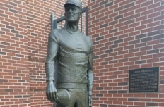 Standing figure of John Heisman, his right hand holding a bullhorn resting on his right thigh.
