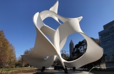 The Koan sculpture by John Portman, bright white undulating ribbons of steel bend in and around eachother, rising up from a granite base. Around the sculpture and through the openings you see a blue sky, tall buildings and trees with autum leaves.
