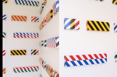 Three white walls, at 90 degree angles to each other, are hung with more than 30 different flat, rectangular canvas each covered in different combinations of industrial safety tape.