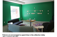 In a room with a bright window and dark green walls there are postcards affixed to the wall and couches and chairs arrayed facing the wall.