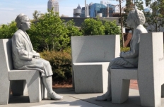 Figures of Rosa Parks at age 42 and age 92, seated on separate chairs facing eachother with an empty chair between them.