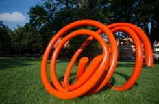 Bright orange steel tubing in a twisted circular pattern on its side in the grass.