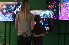 A woman and a child stand with their backs to the camera, engrossed in the images on the monitors before them