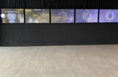 Five monitors are hung next to eachother on a black curtain. Each displays a visual representation of adifferent set of data 