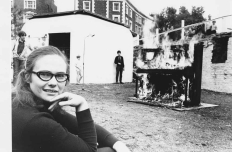 A black and white image from 1968 of a woman smiling at the camera, an upright grand piano in flames in the background