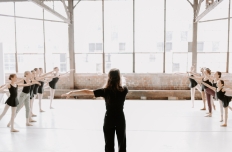 A woman facing a wall of windows teaching a ballet class filled with young dancers