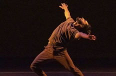 a dancer in all brown, lunging forward with his chest thrust forward