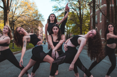 Six white women in black crop tops and leggings in various dance poses in the middle of an empty street