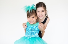 A young dancer in black leotard leaning over the shoulder of a young girl in bright teal sparkly dress and headband
