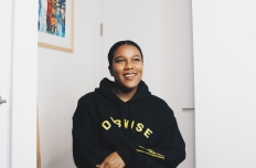 A woman with her hair tied back, in a black hoody with yellow text on the chest and forearm areas, folds her arms as she looks up at the top right corner into the distance. She is in an angled small room painted white, with a colorful framed photo on the left wall.  