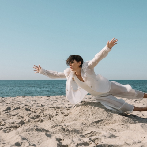 A woman with short dark hair, wearing a white, loose-fitting suit, is on the beach, the blue-grey ocean behind her and the pale grey sand below her. She is nearly horizontal to the sand, as if flying or falling.