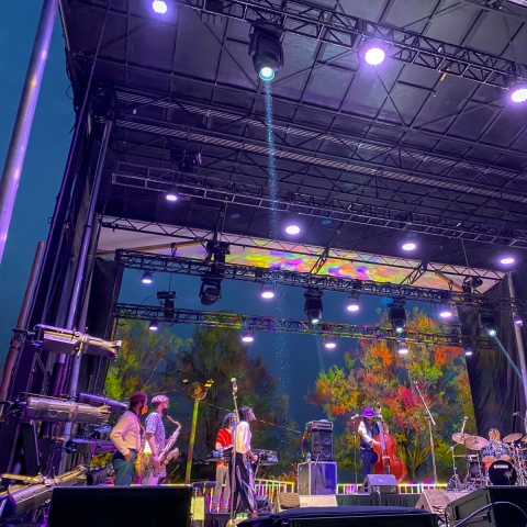 An outdoor stage truss frames musicians who are lit by the multi colored lights that also are projected onto the trees behind them