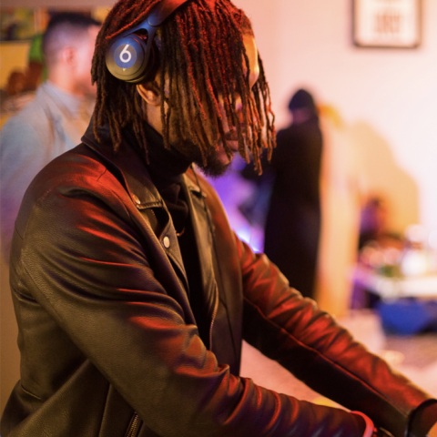 A Dj with short dreads wears Beats Headphones and a black leather jacket as he adjusts a DJ table set, his laptop computer propped up on a stand in front of him, displaying different sound samples. Behind it is a cactus plant. The blurred background behind him is a room with two other people whose faces cannot be seen. In the room is a table with items on top, and a couple of lamps, a picture frame.