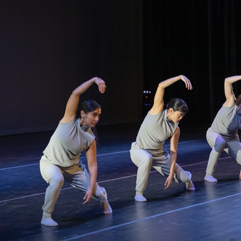 five dancers on stage wearing light grey shirt and pants with one arm curved above their heads and and body lowered toward ground