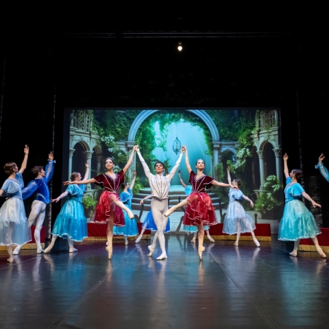 group of ballet dancers on stage wearing blue and red attire with garden scene displayed in background